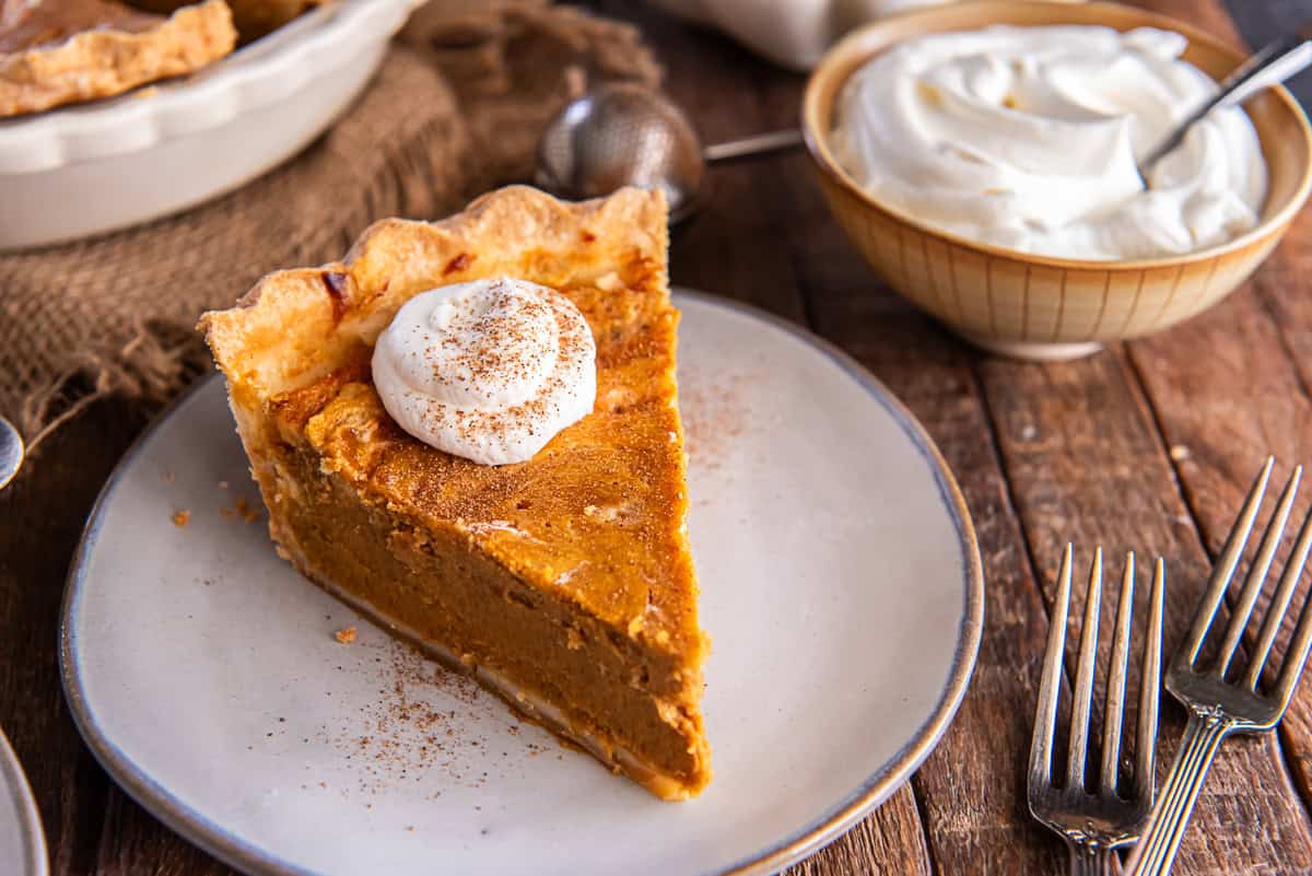 Slice of Hazelnut Pumpkin Pie topped with whipped cream and a sprinkle of cinnamon on a white plate, with a bowl of whipped cream and rustic fall decor in the background.