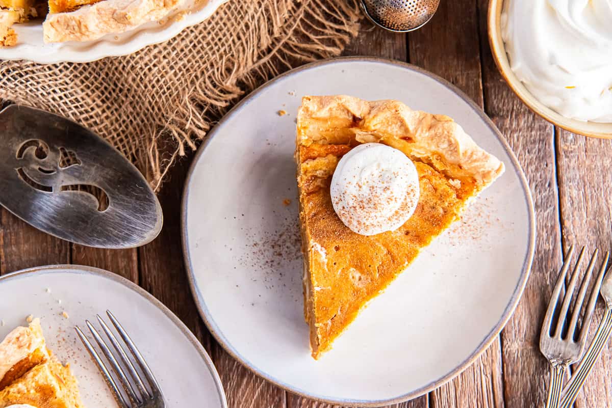 Overhead view of a slice of Hazelnut Pumpkin Pie topped with whipped cream on a plate beside forks and a bowl of whipped cream on a wooden table.
