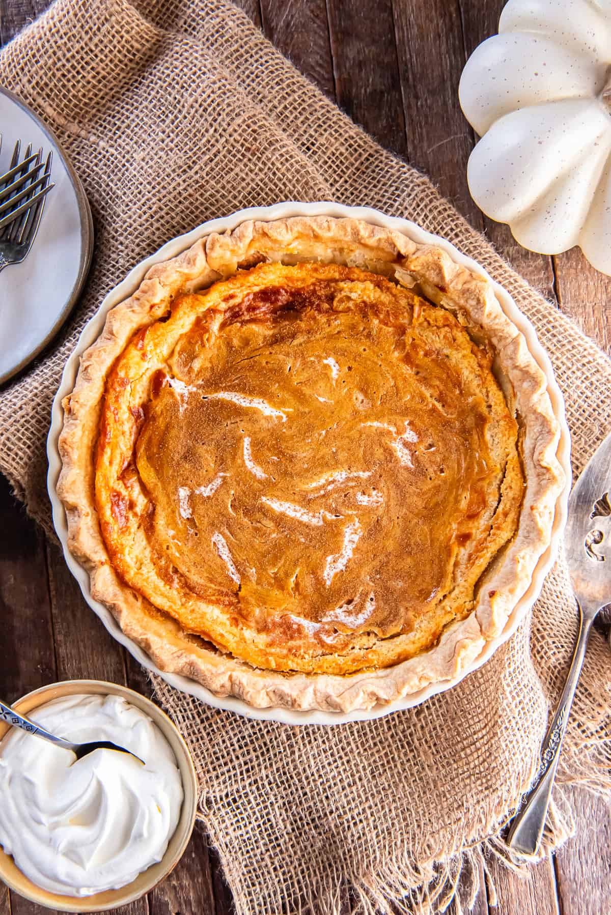 Whole Hazelnut Pumpkin Pie with a golden marbled top in a flaky crust, styled on a rustic burlap surface with whipped cream and serving utensils nearby.