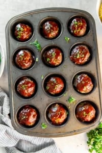 Muffin pan filled with glazed meatloaf muffins surrounded by fresh parsley