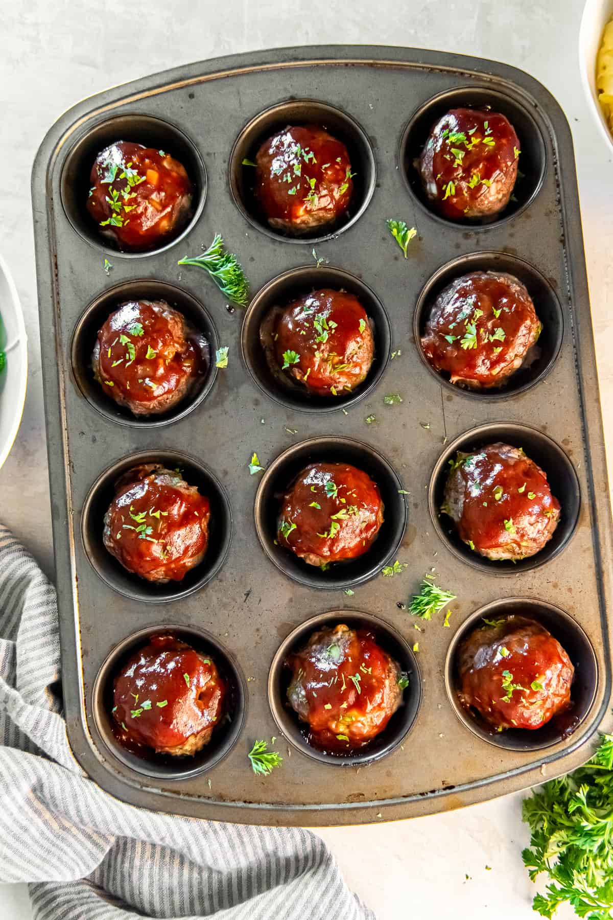 Muffin pan filled with glazed meatloaf muffins surrounded by fresh parsley
