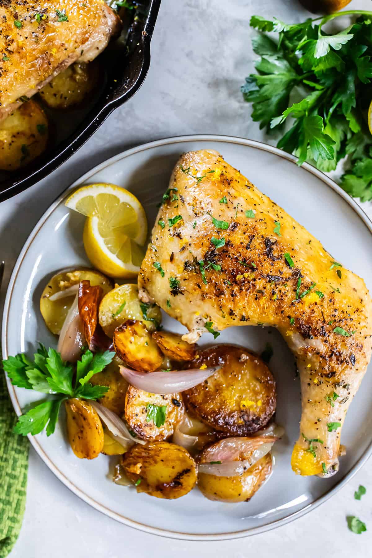 A roasted chicken leg quarter on a white plate with lemon wedges and potatoes next to a cast iron skillet.