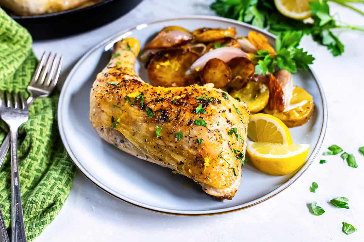 A roasted chicken leg quarter on a white plate with lemon wedges and potatoes.
