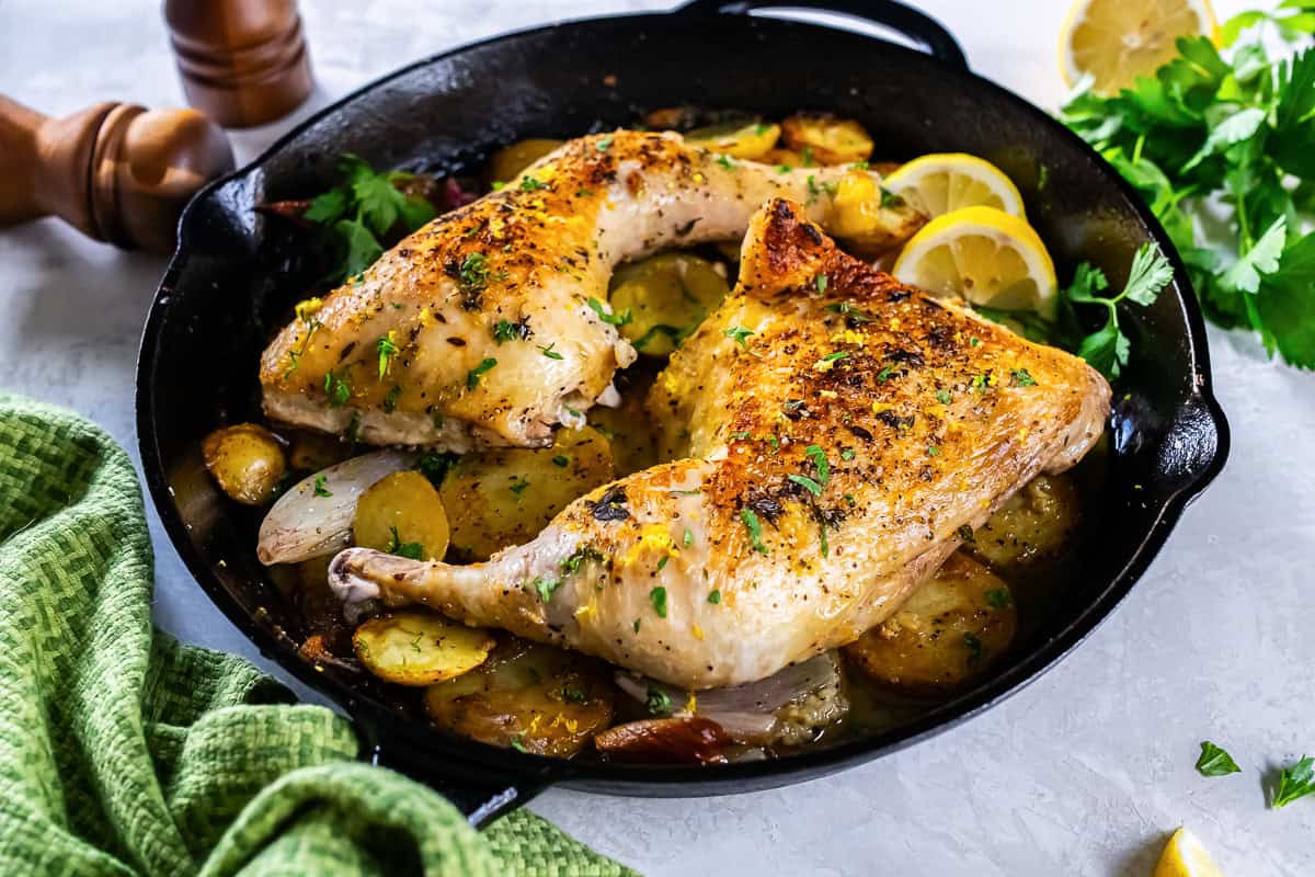 Crispy roasted chicken leg quarters with potatoes and lemon in a cast-iron pan.