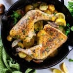 Roasted chicken leg quarters in a cast-iron skillet with potatoes, lemon wedges, and herbs.