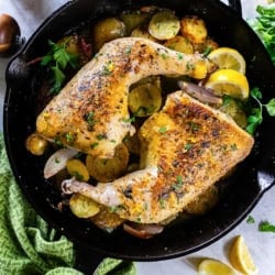 Roasted chicken leg quarters in a cast-iron skillet with potatoes, lemon wedges, and herbs.