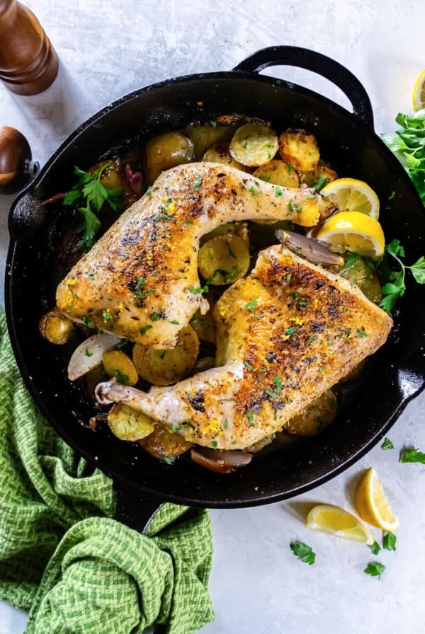 Roasted chicken leg quarters in a cast-iron skillet with potatoes, lemon wedges, and herbs.