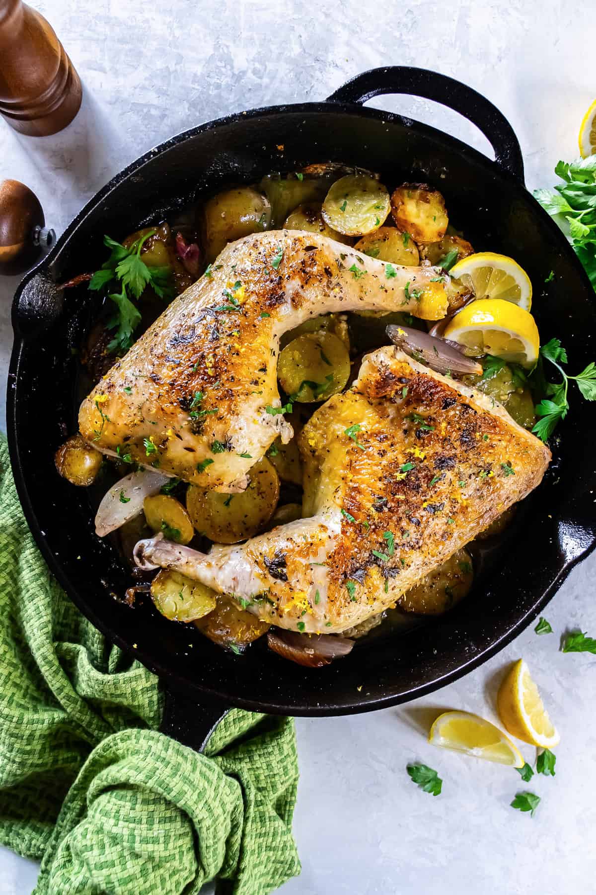 Roasted chicken leg quarters in a cast-iron skillet with potatoes, lemon wedges, and herbs.