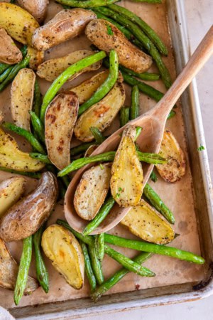 A wooden spoon resting on a sheet pan full of roasted potatoes and green beans.