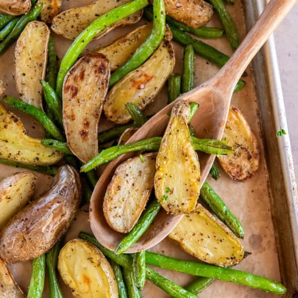 A wooden spoon resting on a sheet pan full of roasted potatoes and green beans.