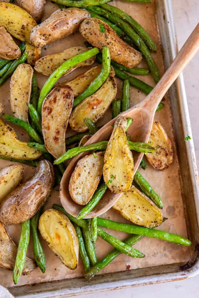 A wooden spoon resting on a sheet pan full of roasted potatoes and green beans.