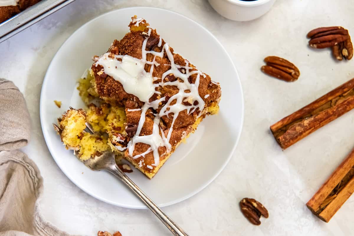 Slice of coffee cake topped with icing and pecans on a white plate with a fork, surrounded by cinnamon sticks, pecans, and a cup of coffee.