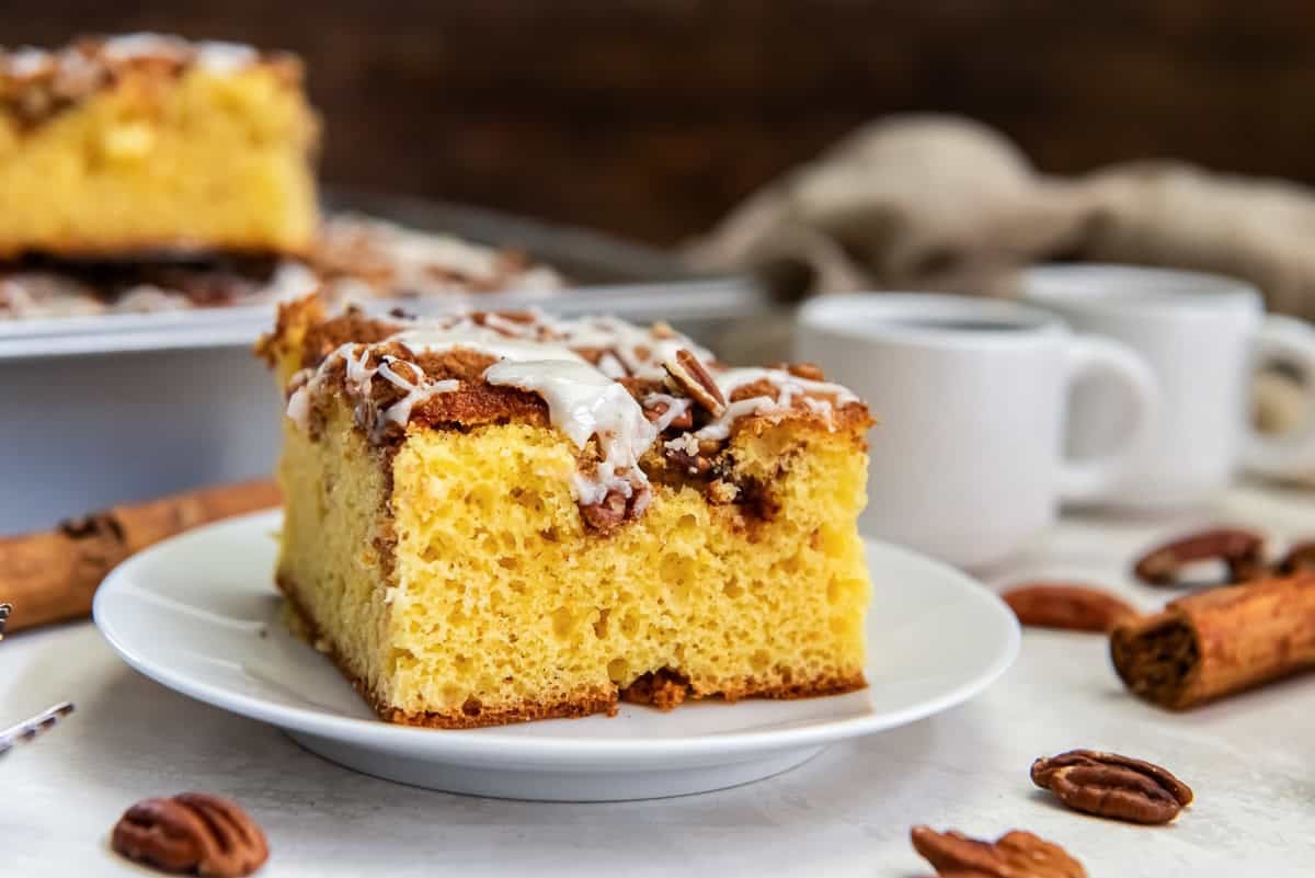 Slice of coffee cake with pecan streusel and icing on a white plate, stacked on dishes with coffee cups and the remaining cake in the background.