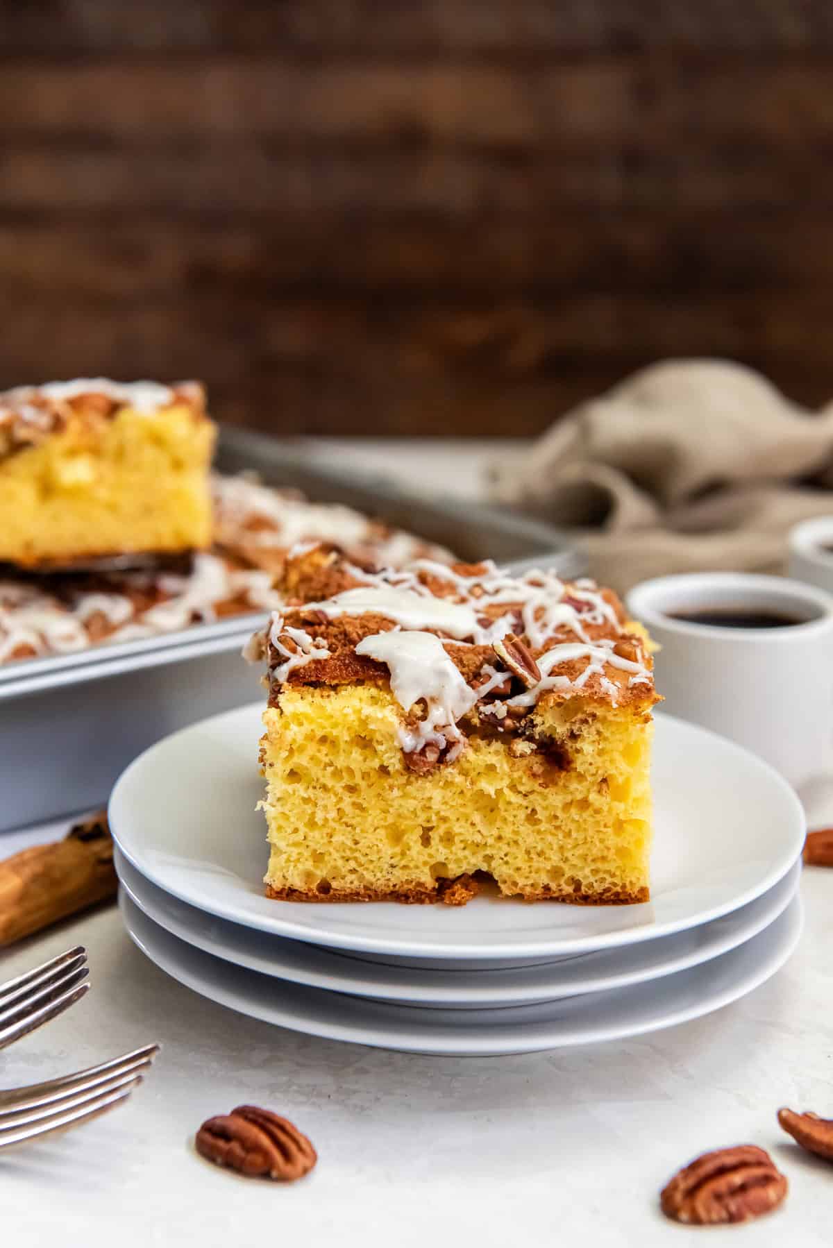 Slice of cake mix coffee cake on a white plate, topped with chopped pecans and a drizzle of icing, with coffee cups and the remaining cake in the background.