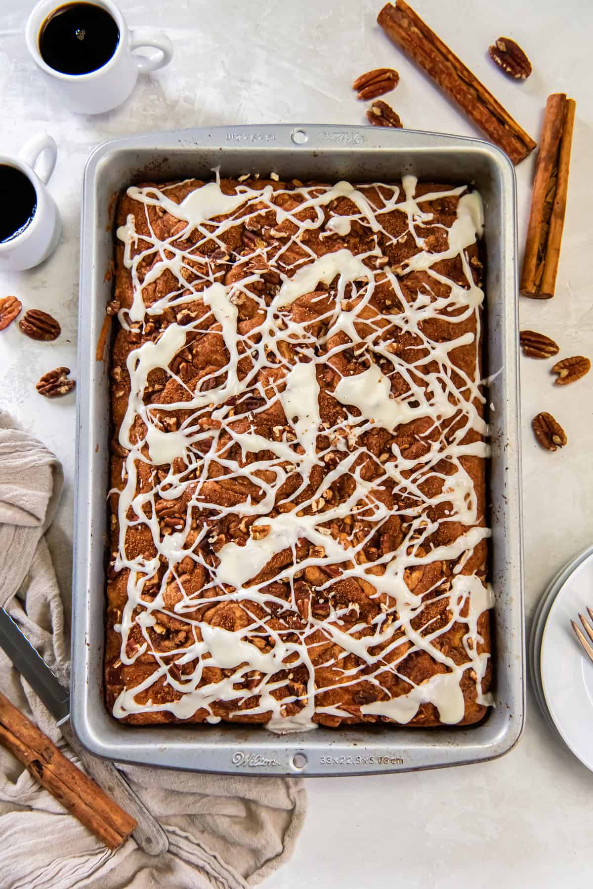 Cake mix coffee cake in a pan topped with chopped pecans and drizzled with white icing, with coffee cups and cinnamon sticks nearby.