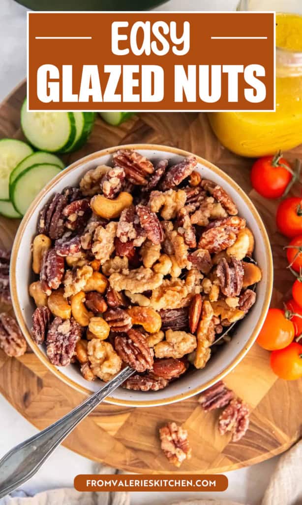 A bowl of glazed mixed nuts with pecans, walnuts, almonds, and cashews on a wooden board with cherry tomatoes, sliced cucumbers, and a jar of yellow vinaigrette in the background with text overlay.