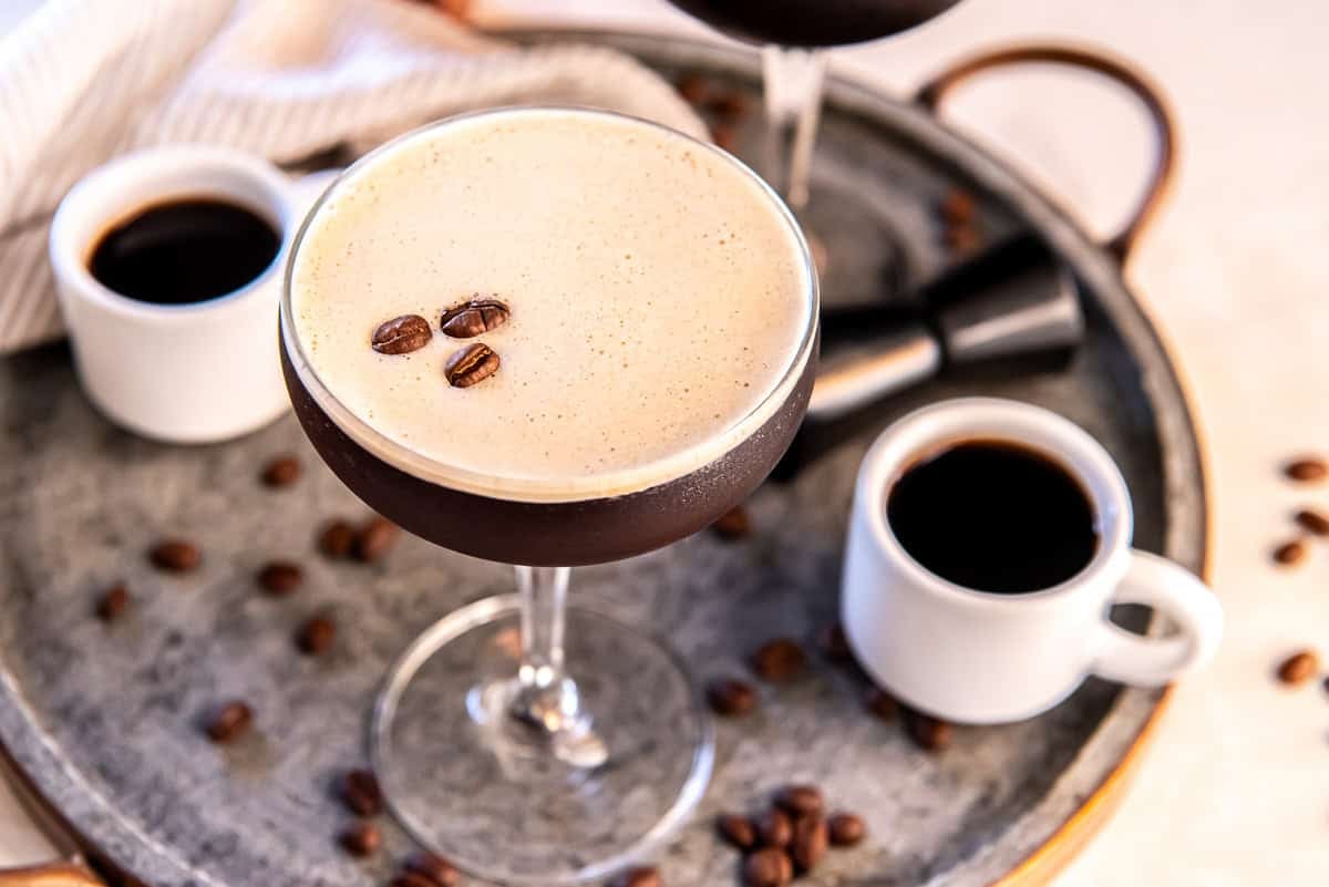 A chilled espresso martini in a coupe glass topped with a thick layer of creamy foam and three coffee beans, set on a round metal tray with scattered coffee beans, two small cups of espresso, and a cocktail jigger.
