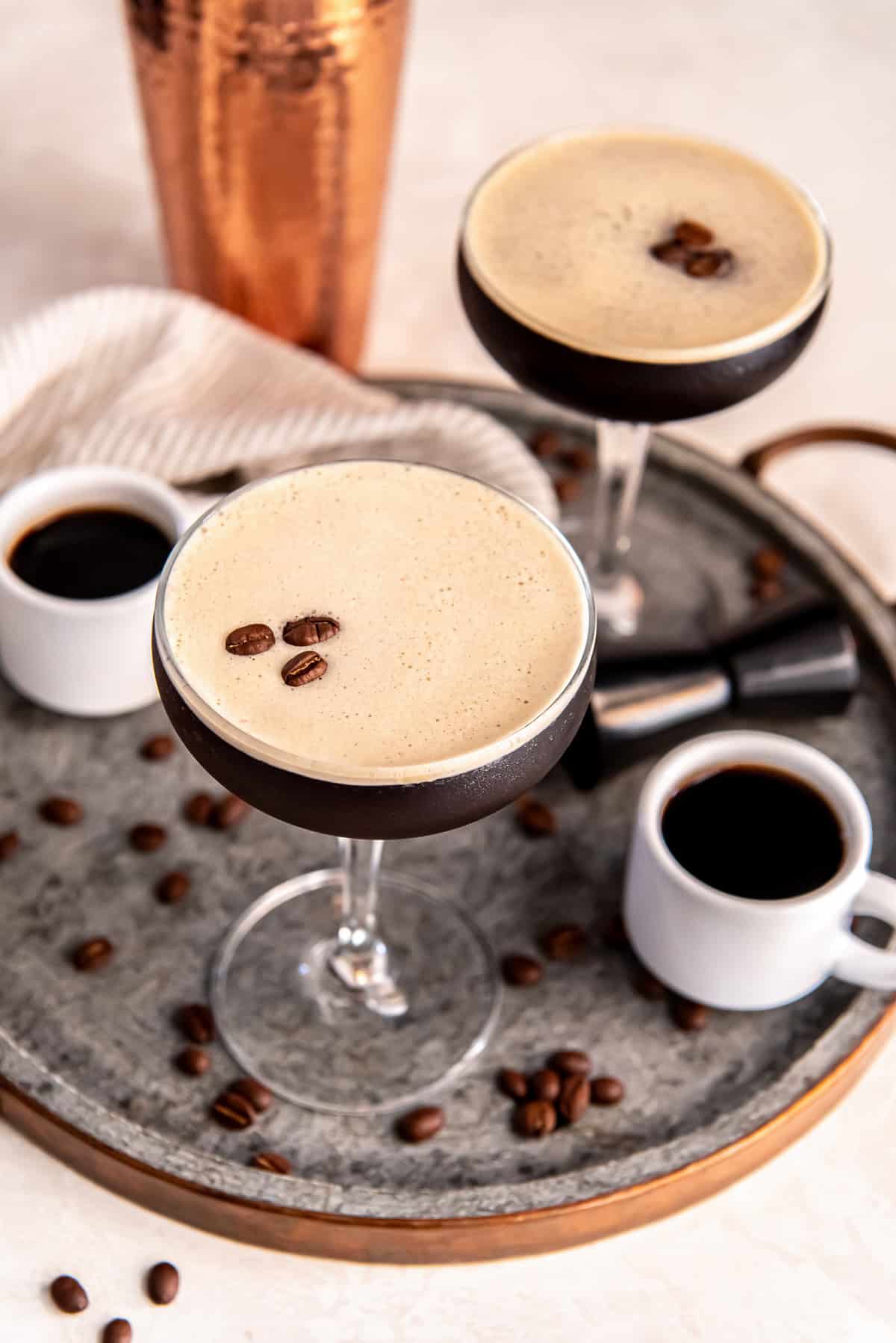 A chilled espresso martini in a coupe glass topped with creamy foam and three coffee beans, set on a round metal tray with scattered coffee beans, espresso cups, and a cocktail jigger.