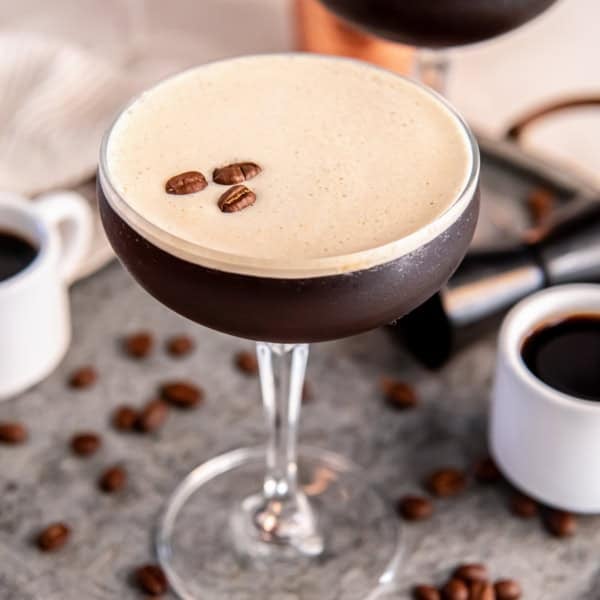 A chilled espresso martini in a coupe glass topped with a thick layer of creamy foam and three coffee beans, set on a metal tray with scattered coffee beans, small espresso cups, and a cocktail jigger in the background.