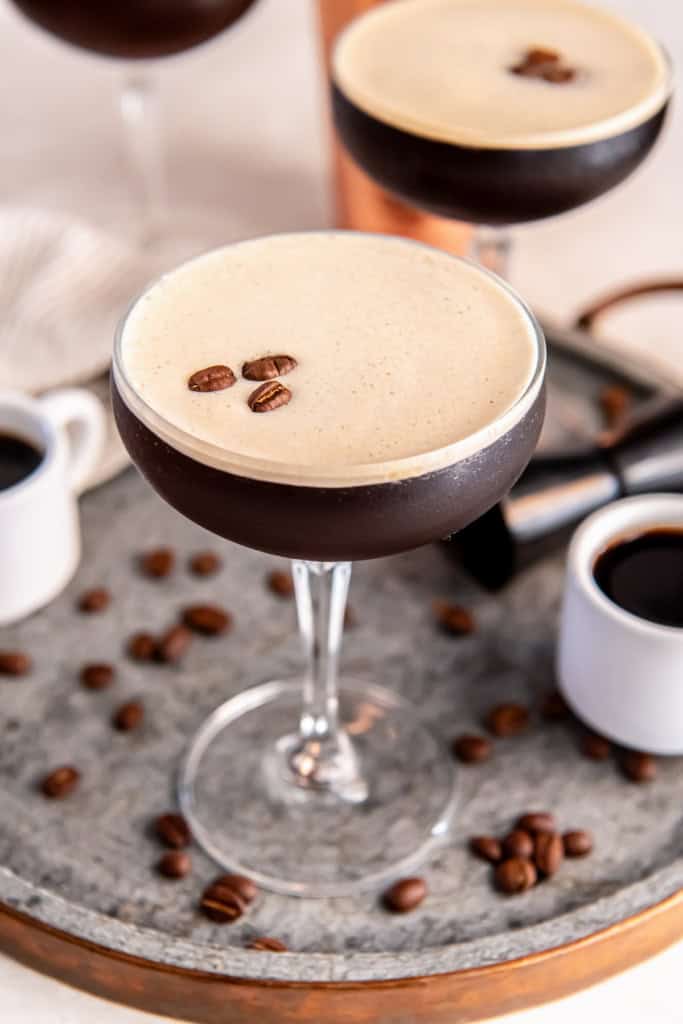 A chilled espresso martini in a coupe glass topped with a thick layer of creamy foam and three coffee beans, set on a metal tray with scattered coffee beans, small espresso cups, and a cocktail jigger in the background.