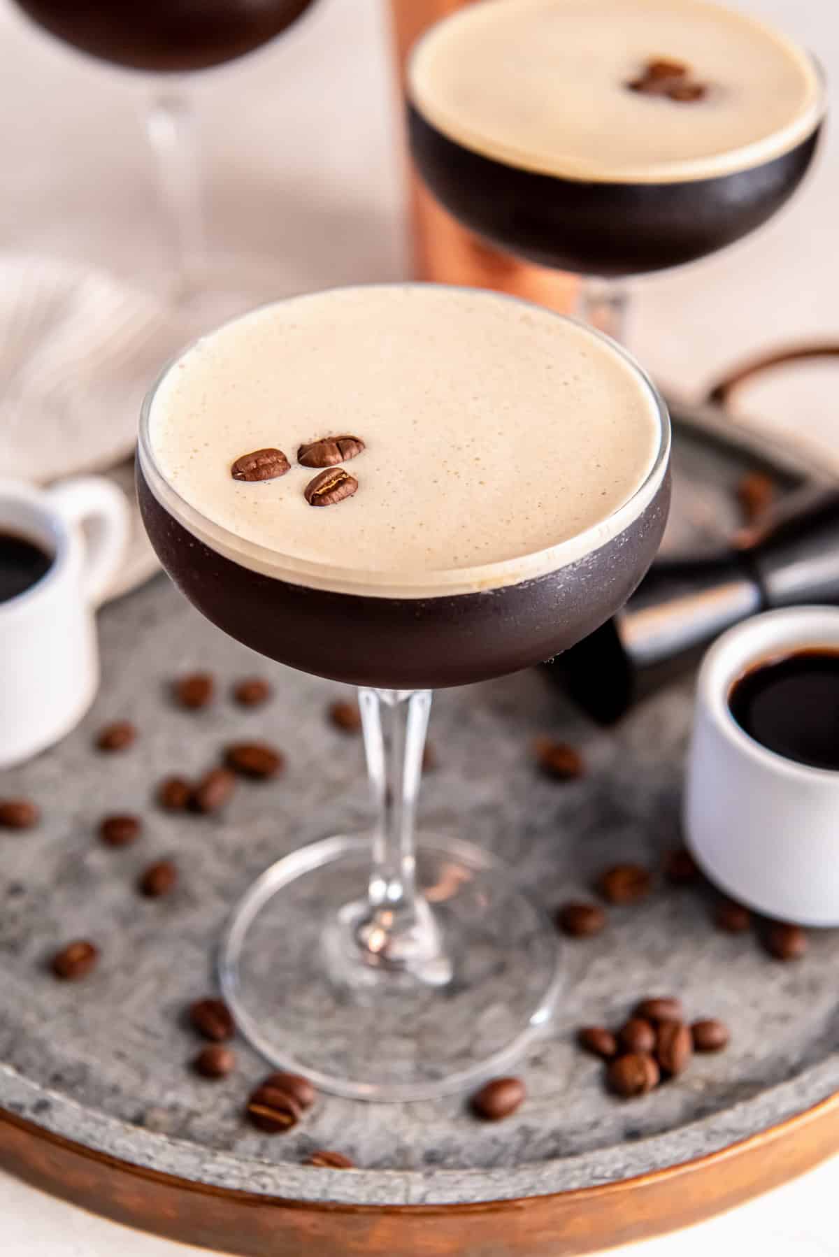 A chilled espresso martini in a coupe glass topped with a thick layer of creamy foam and three coffee beans, set on a metal tray with scattered coffee beans, small espresso cups, and a cocktail jigger in the background.