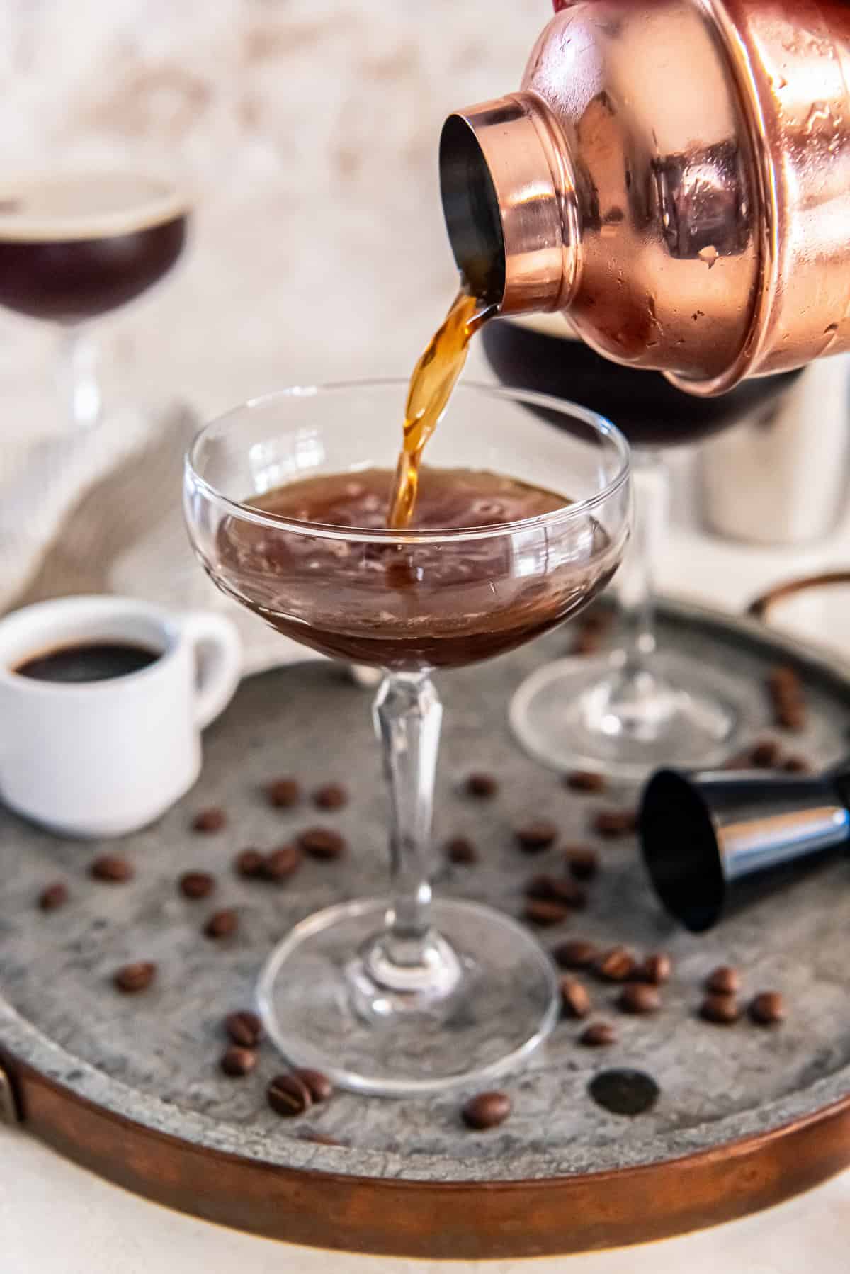 A copper cocktail shaker pours an espresso martini mixture into a coupe glass on a metal tray scattered with coffee beans, with a small cup of espresso and a second cocktail glass in the background.