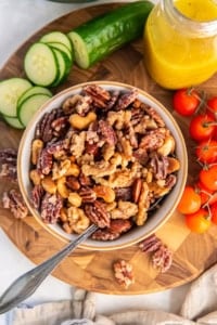 A bowl of glazed mixed nuts with pecans, walnuts, almonds, and cashews on a wooden board with cherry tomatoes, sliced cucumbers, and a jar of yellow vinaigrette in the background.