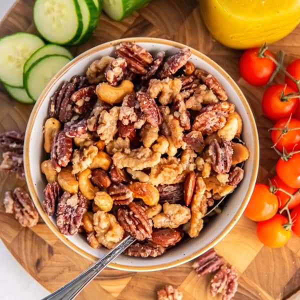 A bowl of glazed mixed nuts with pecans, walnuts, almonds, and cashews on a wooden board with cherry tomatoes, sliced cucumbers, and a jar of yellow vinaigrette in the background.
