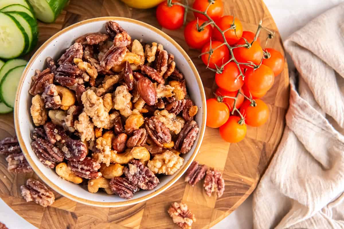 A bowl of glazed mixed nuts with pecans, walnuts, almonds, and cashews on a wooden board surrounded by cherry tomatoes, sliced cucumbers, and a beige linen napkin.