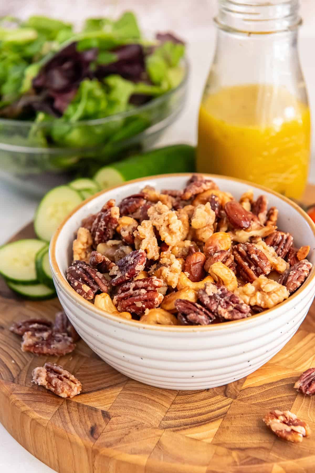 A white bowl filled with glazed mixed nuts including pecans, walnuts, almonds, and cashews, set on a wooden board with salad greens and a jar of dressing in the background.