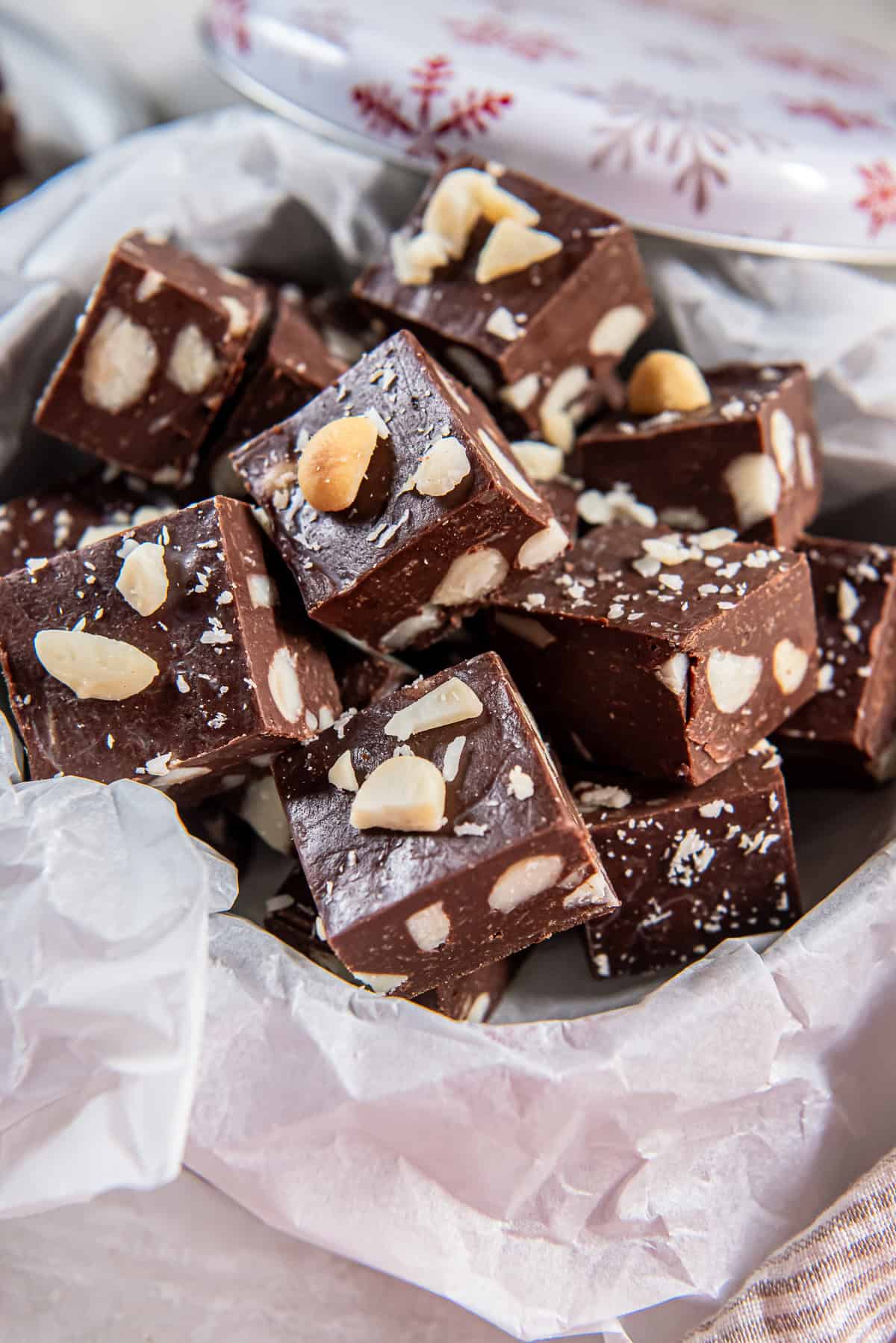Macadamia nut fudge in a parchment-lined holiday tin with the lid resting behind it.