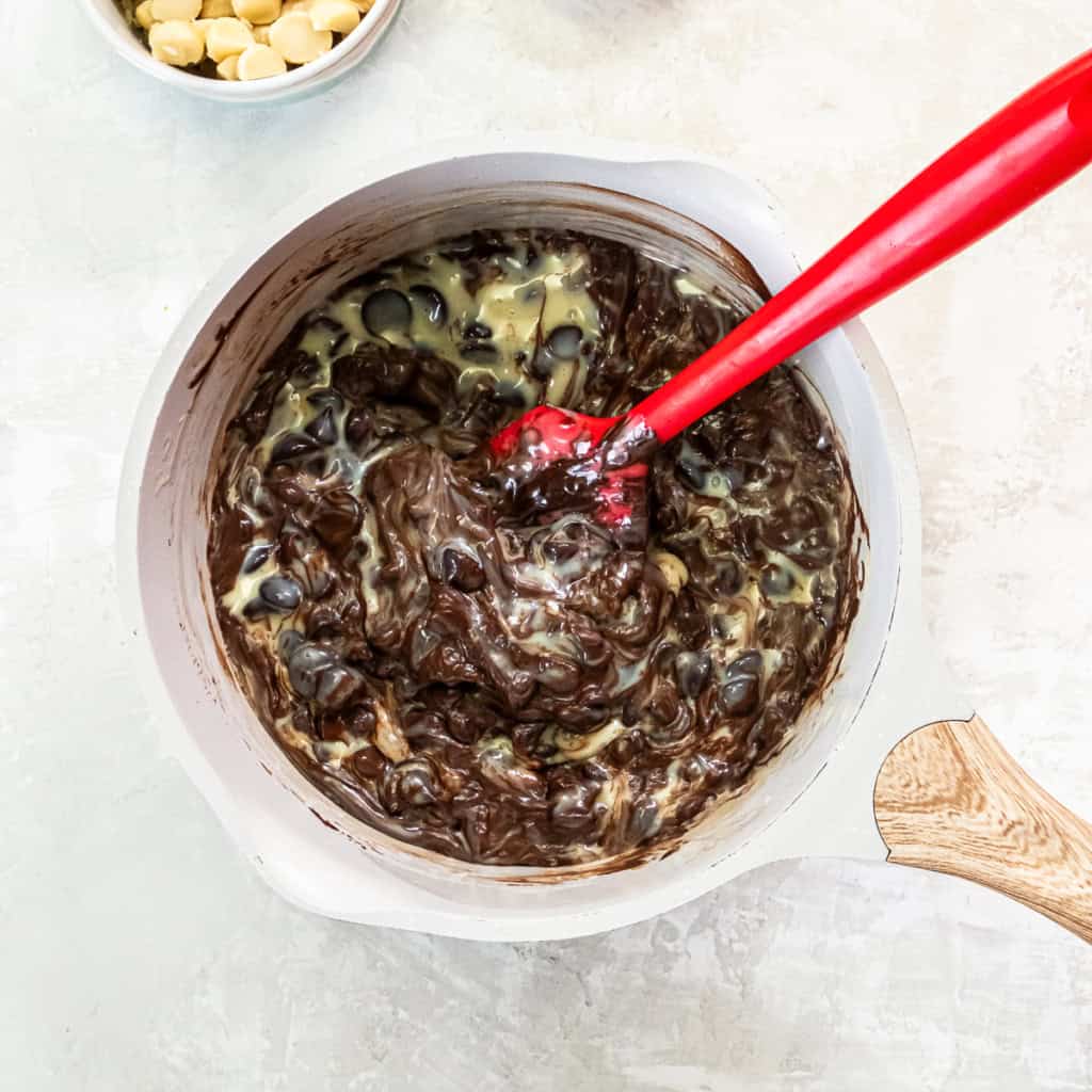 A red spoon stirring melted chocolate chips and sweetened condensed milk in a saucepan.