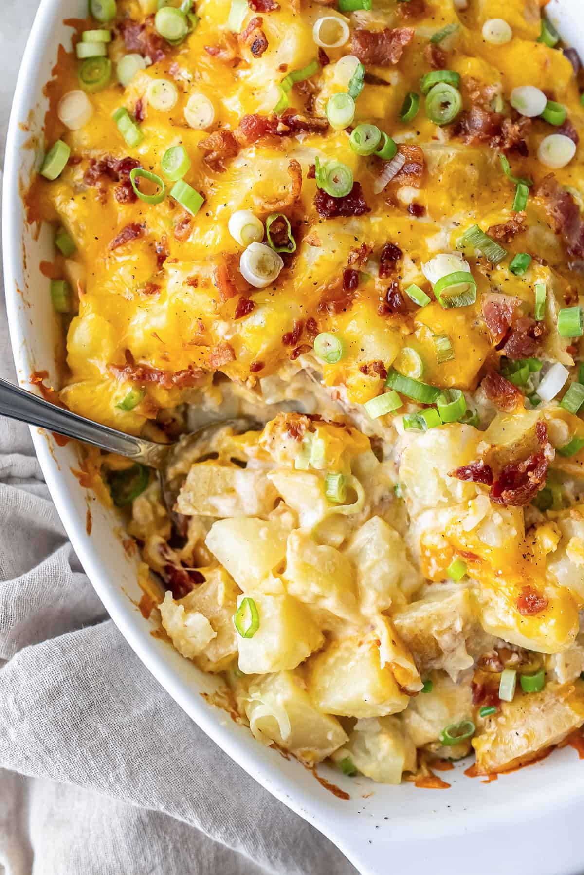Twice Baked Potato Casserole in a white baking dish with melted cheddar cheese, crispy bacon, and sliced green onions on top. A spoon is scooping out a creamy serving of cheesy potatoes.