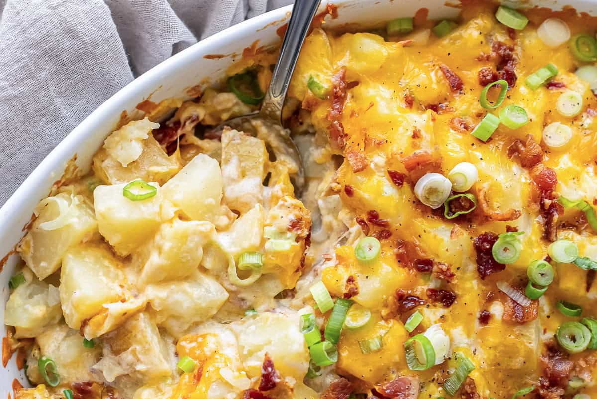 A spoon resting in a white baking dish full of Twice Baked Potato Casserole with cheese and bacon.
