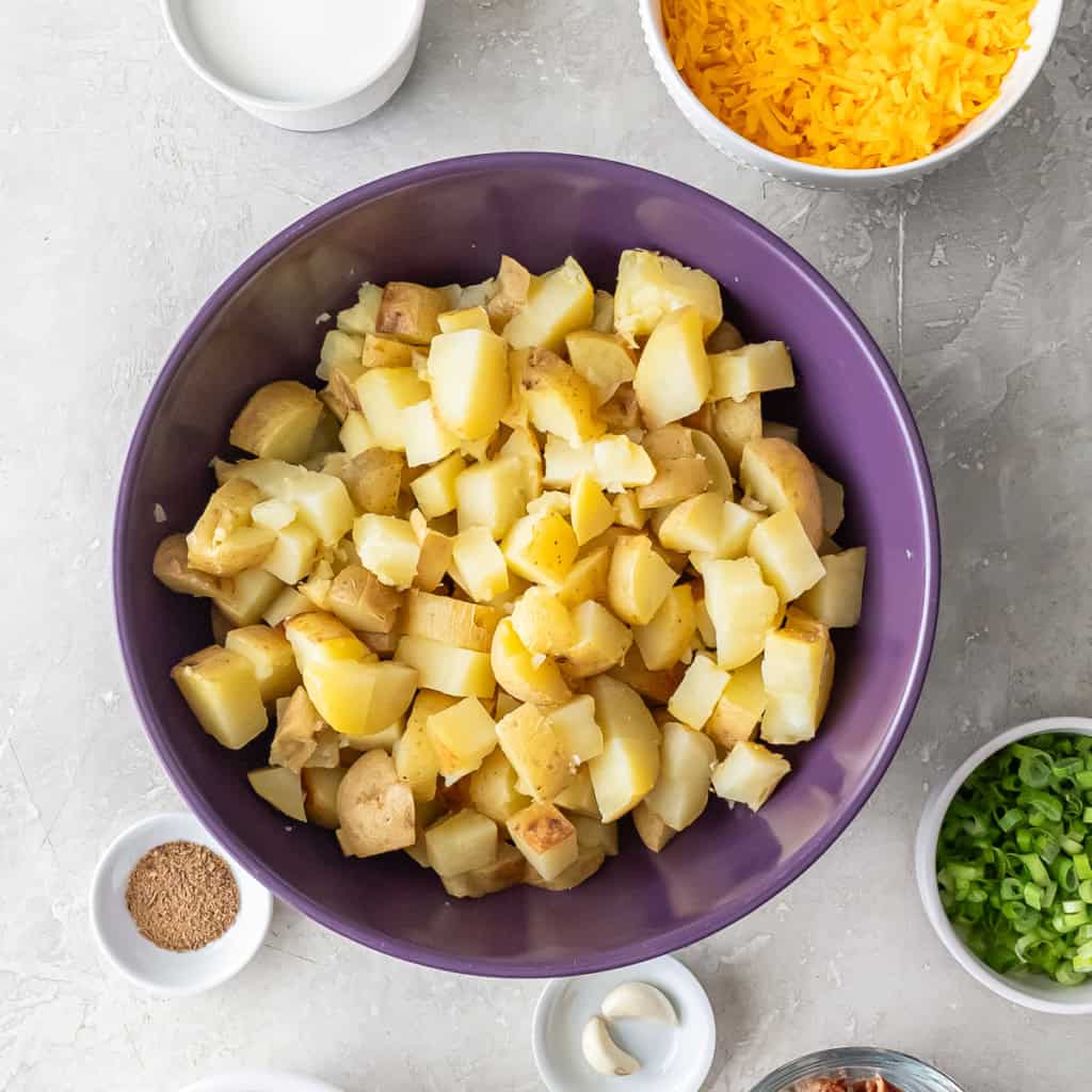 Bowl of chopped baked Yukon Gold potatoes in a large purple mixing bowl with milk, cheese, seasoning, and other ingredients arranged around it.