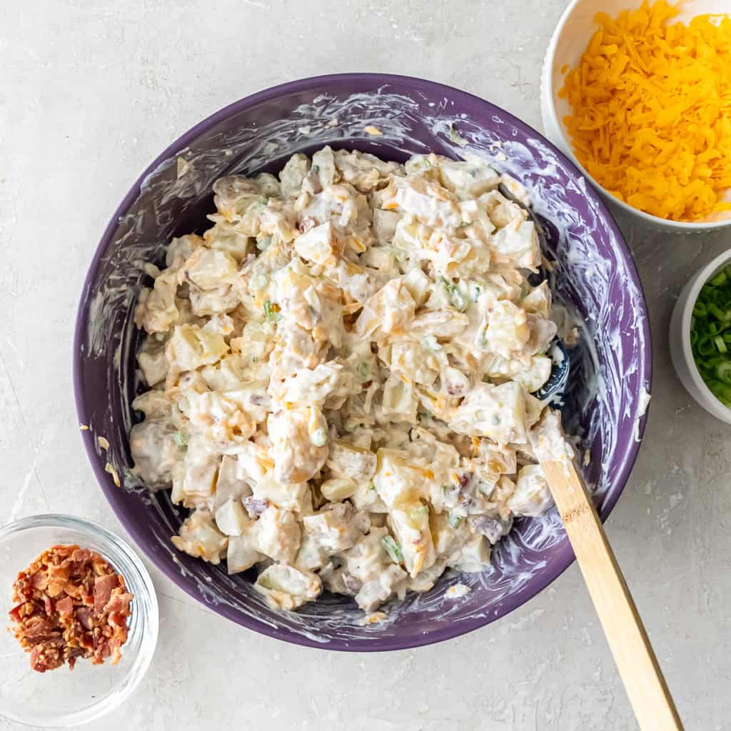 Potato mixture combined with sour cream, cheese, green onions, and bacon in a purple mixing bowl.
