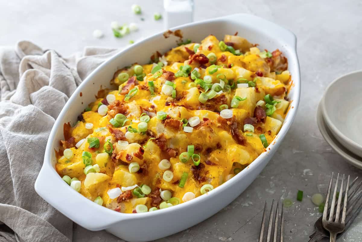 Twice Baked Potato Casserole in a white baking dish with melted cheddar cheese, bacon, and green onions, set on a neutral surface with a linen napkin and forks beside it.