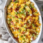 wice Baked Potato Casserole in a white baking dish topped with melted cheddar, crisp bacon, and green onions, set on a neutral linen with forks and plates nearby.