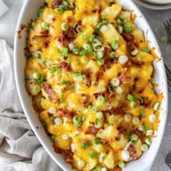 wice Baked Potato Casserole in a white baking dish topped with melted cheddar, crisp bacon, and green onions, set on a neutral linen with forks and plates nearby.