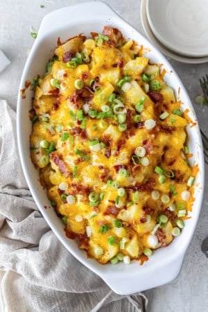 wice Baked Potato Casserole in a white baking dish topped with melted cheddar, crisp bacon, and green onions, set on a neutral linen with forks and plates nearby.