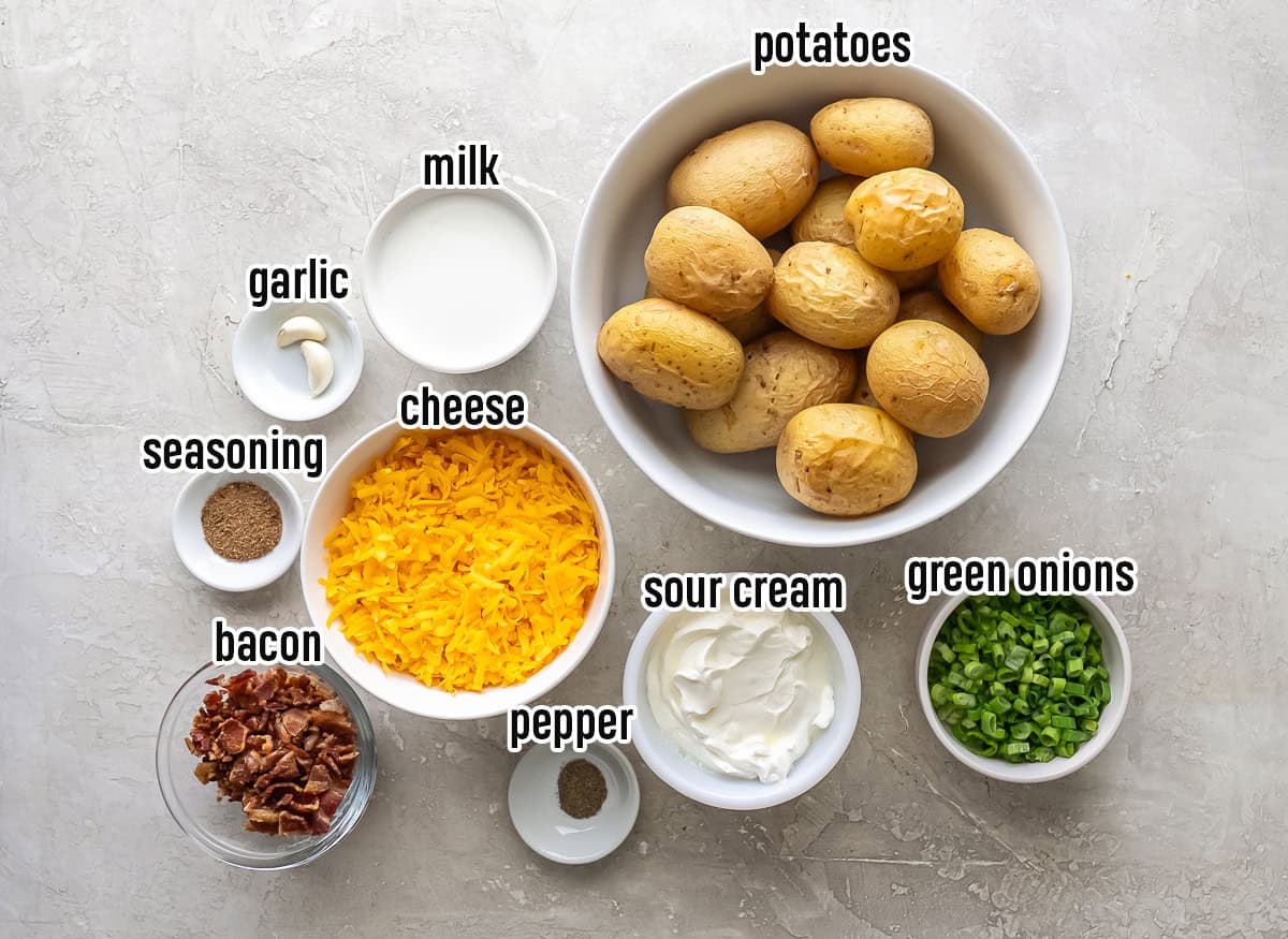 ingredients for Twice Baked Potato Casserole, including whole baked Yukon Gold potatoes, shredded cheddar cheese, sour cream, milk, garlic, seasoning, black pepper, crisp cooked bacon, and chopped green onions arranged in white bowls on a light surface.
