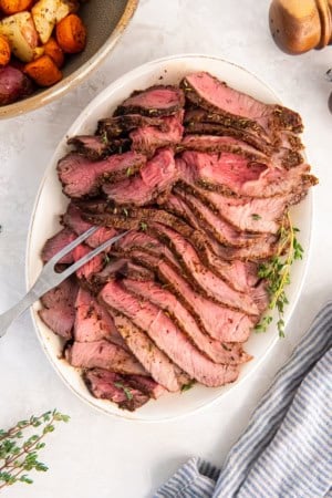 Platter of sliced air fryer sirloin tip roast served medium-rare, with roasted vegetables in the background.