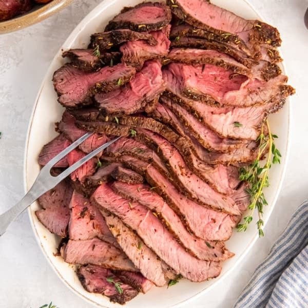 Platter of sliced air fryer sirloin tip roast served medium-rare, with roasted vegetables in the background.