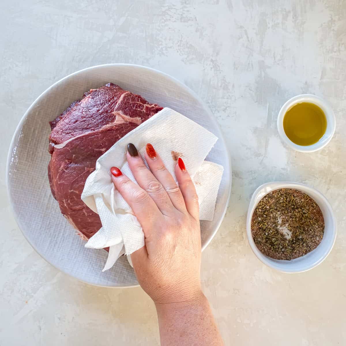 Patting a sirloin tip roast dry with paper towels before seasoning, with olive oil and seasoning mix nearby.