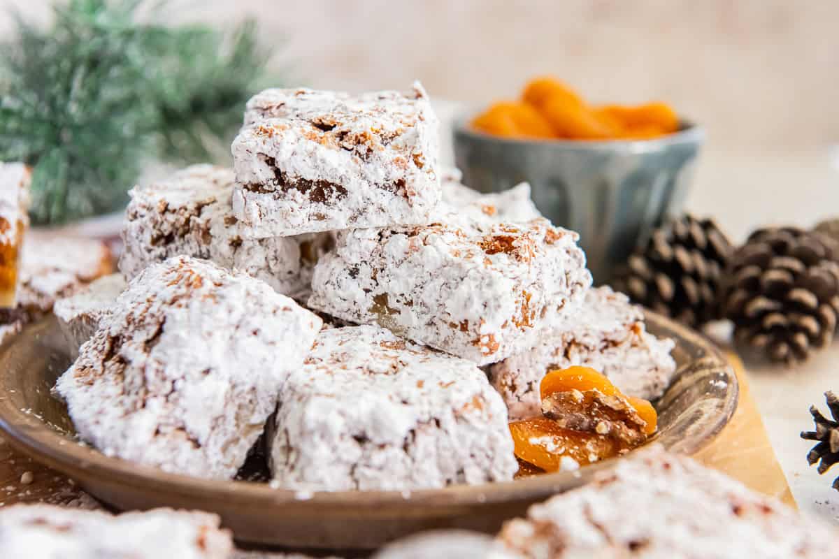 Apricot bars dusted with powdered sugar, stacked on a rustic plate with dried apricots in the background.