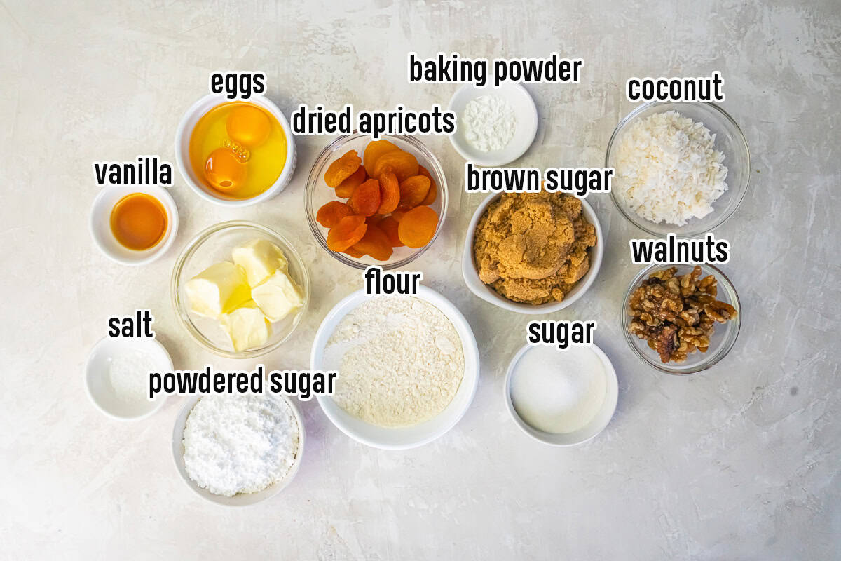 Ingredients for apricot bars, including dried apricots, flour, butter, eggs, brown sugar, walnuts, coconut, and powdered sugar.