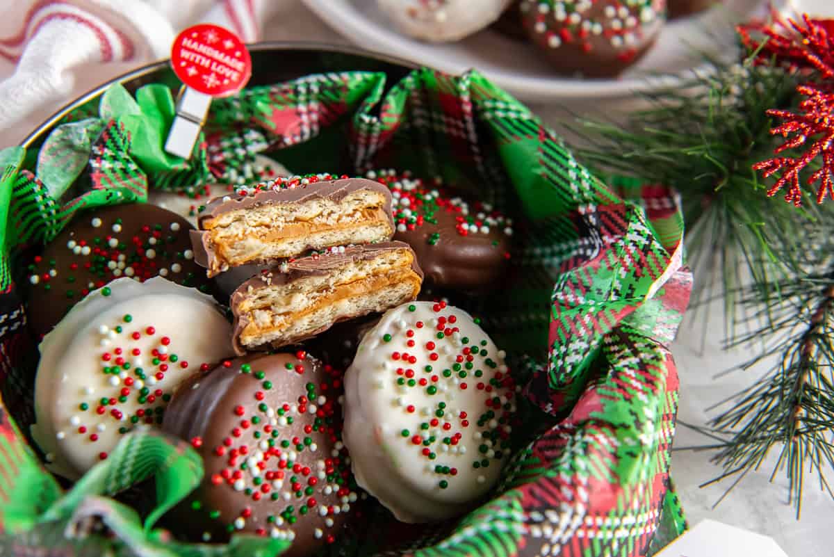 Holiday tin lined with green plaid tissue paper filled with chocolate-dipped peanut butter Ritz cookies, including a stacked cookie cut in half to show the creamy peanut butter center, all topped with festive red, white, and green sprinkles.