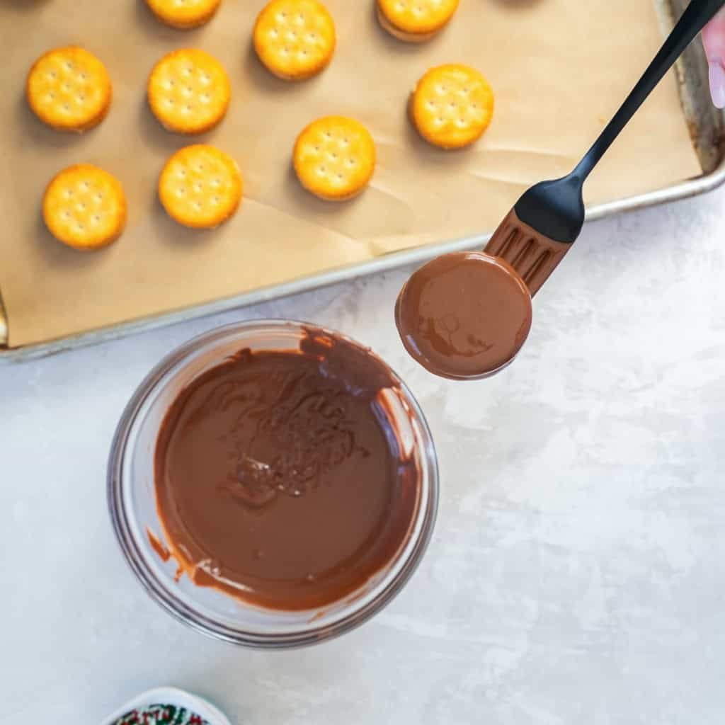 A peanut butter Ritz sandwich cookie coated in melted chocolate held over a bowl of chocolate with undipped cookies on a tray in the background.