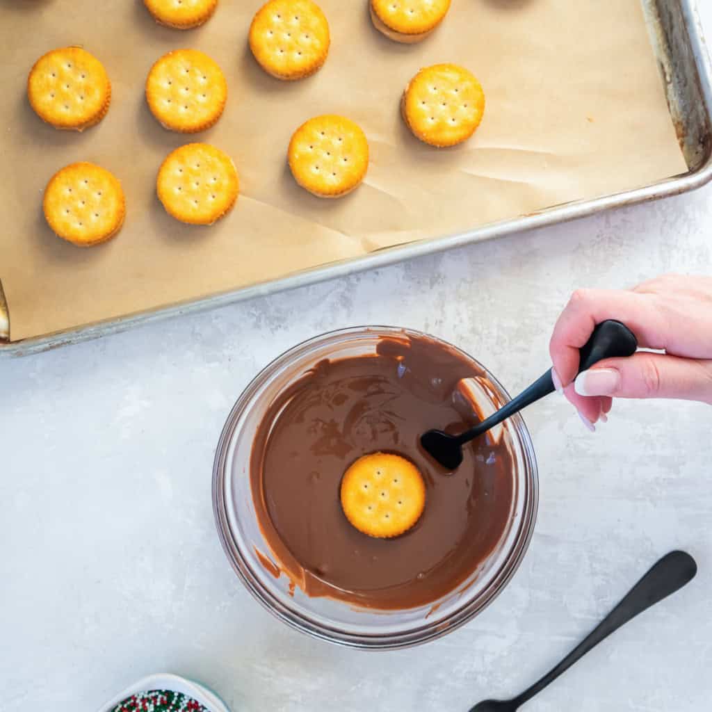 A peanut butter Ritz sandwich cookie being dipped into a bowl of melted chocolate using a fork, with assembled cookies on a parchment-lined tray.