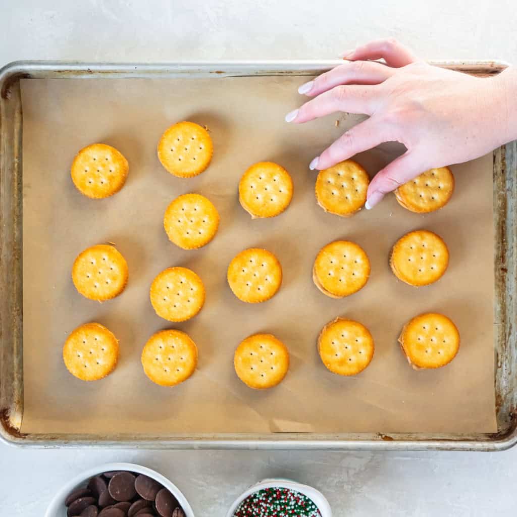 A hand placing a Ritz cracker on top of peanut butter to assemble sandwich cookies on a parchment-lined baking sheet.